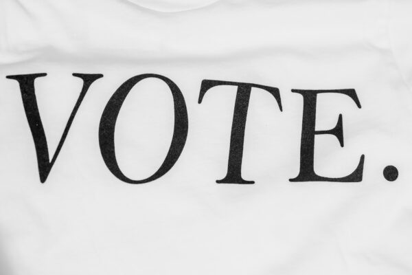 A photo of a white textured background with black text that reads "Vote". Photo was taken by Cyrus Crossan