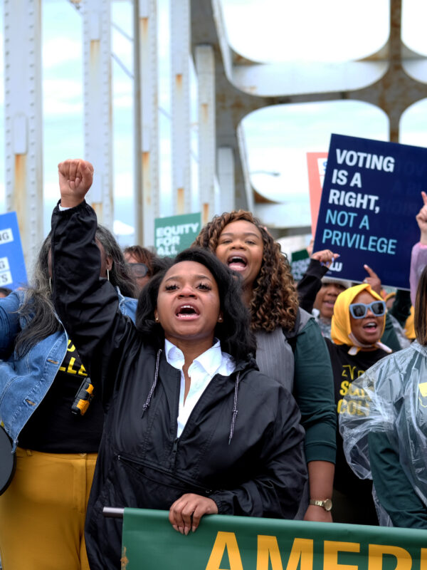 crowd marches with signs behind a banner while marching over the edmund pettus bridge