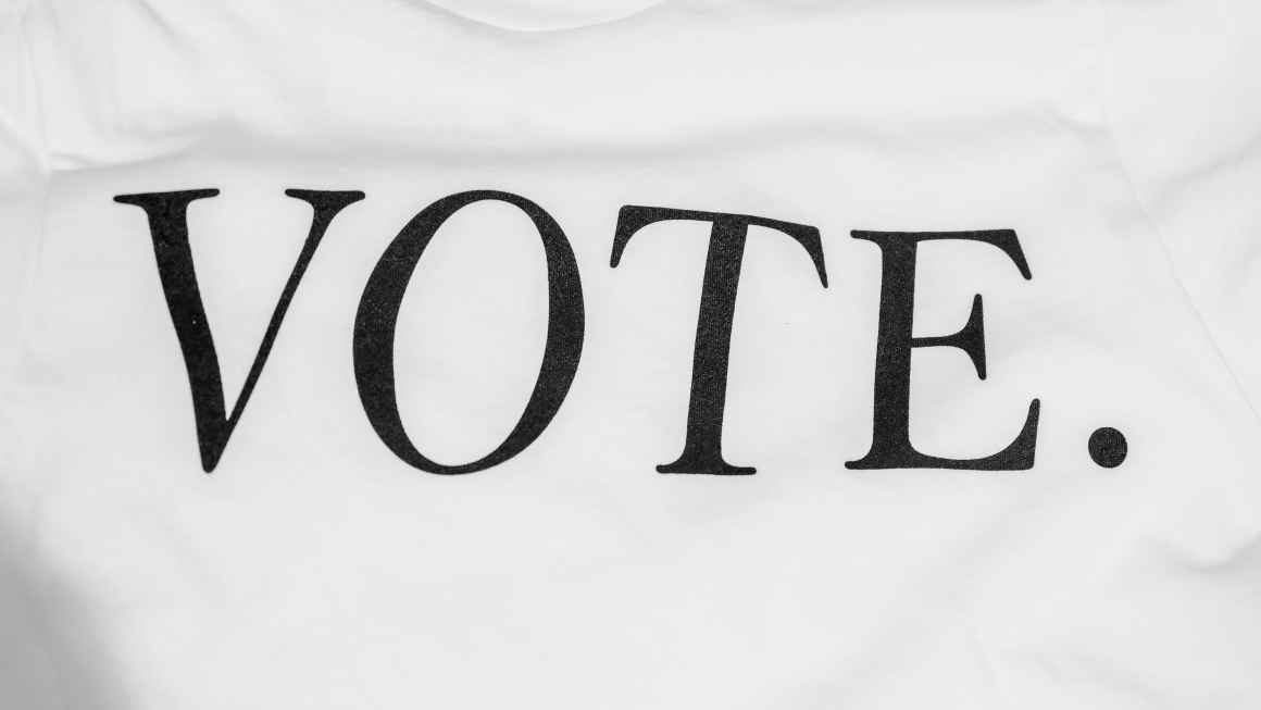 A photo of a white textured background with black text that reads "Vote". Photo was taken by Cyrus Crossan