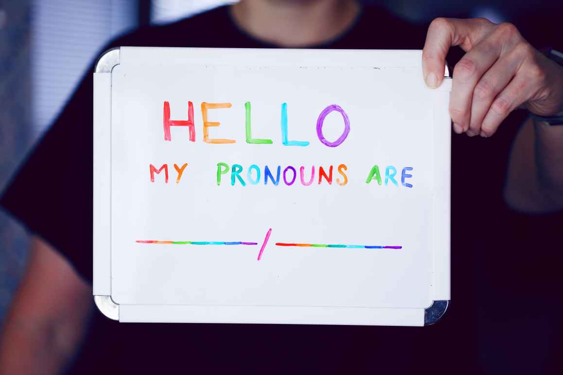 A photo of a person holding up a whiteboard with the words written 'Hello, my pronouns are [blank/blank]. The words are written in rainbow colors.