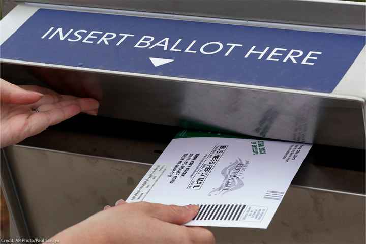 A voter holds the door of a ballot box open with her left hand while inserting her voter ballot with her right hand.